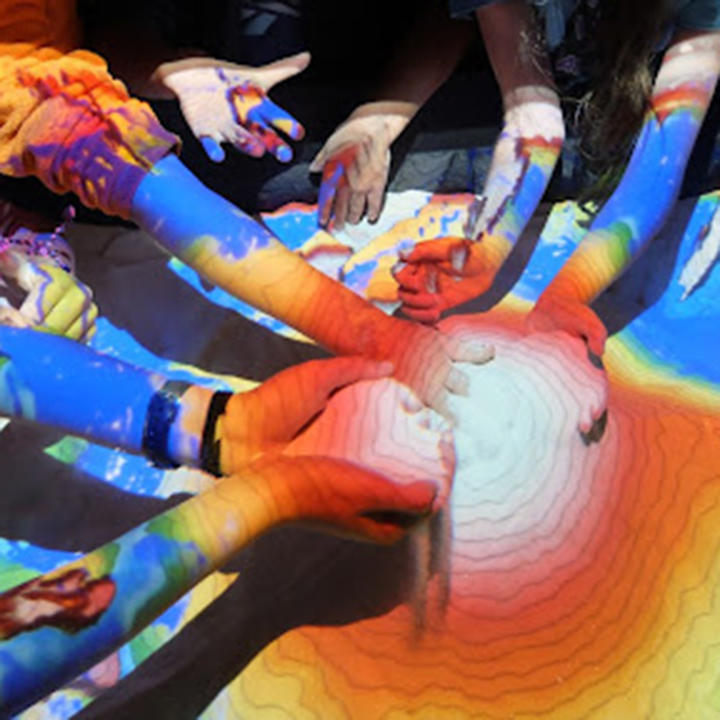 Numerous hands reaching into a sandbox with an AR topographical projection