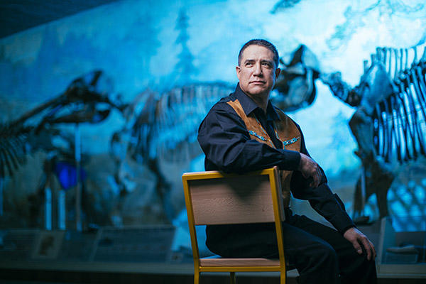 Speaker Lawrence Bradley sitting on a chair, looking over his shoulder in front of dinosaur fossils.
