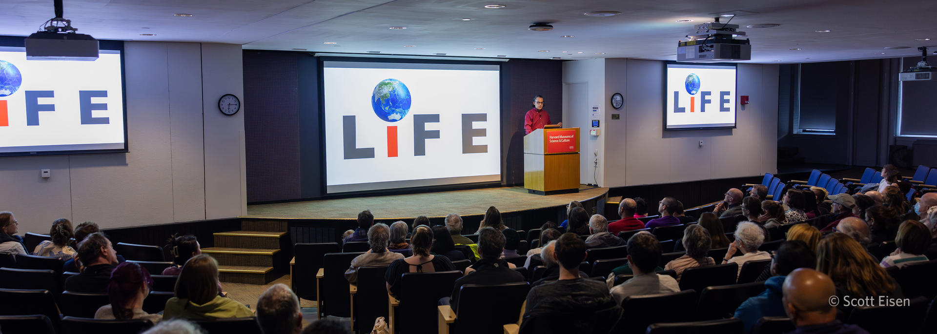 auditorium with speaker at podium in front of 3 large screens reading "life".
